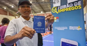 Trabalhador ao lado de um baner sobre feirão de emprego e uma carteira de trabalho em mãos