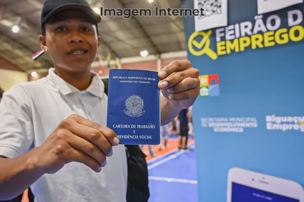 Trabalhador ao lado de um baner sobre feirão de emprego e uma carteira de trabalho em mãos