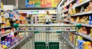 Carrinho de supermercado em corredor com prateleiras cheias de alimentos e preços ao fundo, representando inflação e aumento do custo de vida.